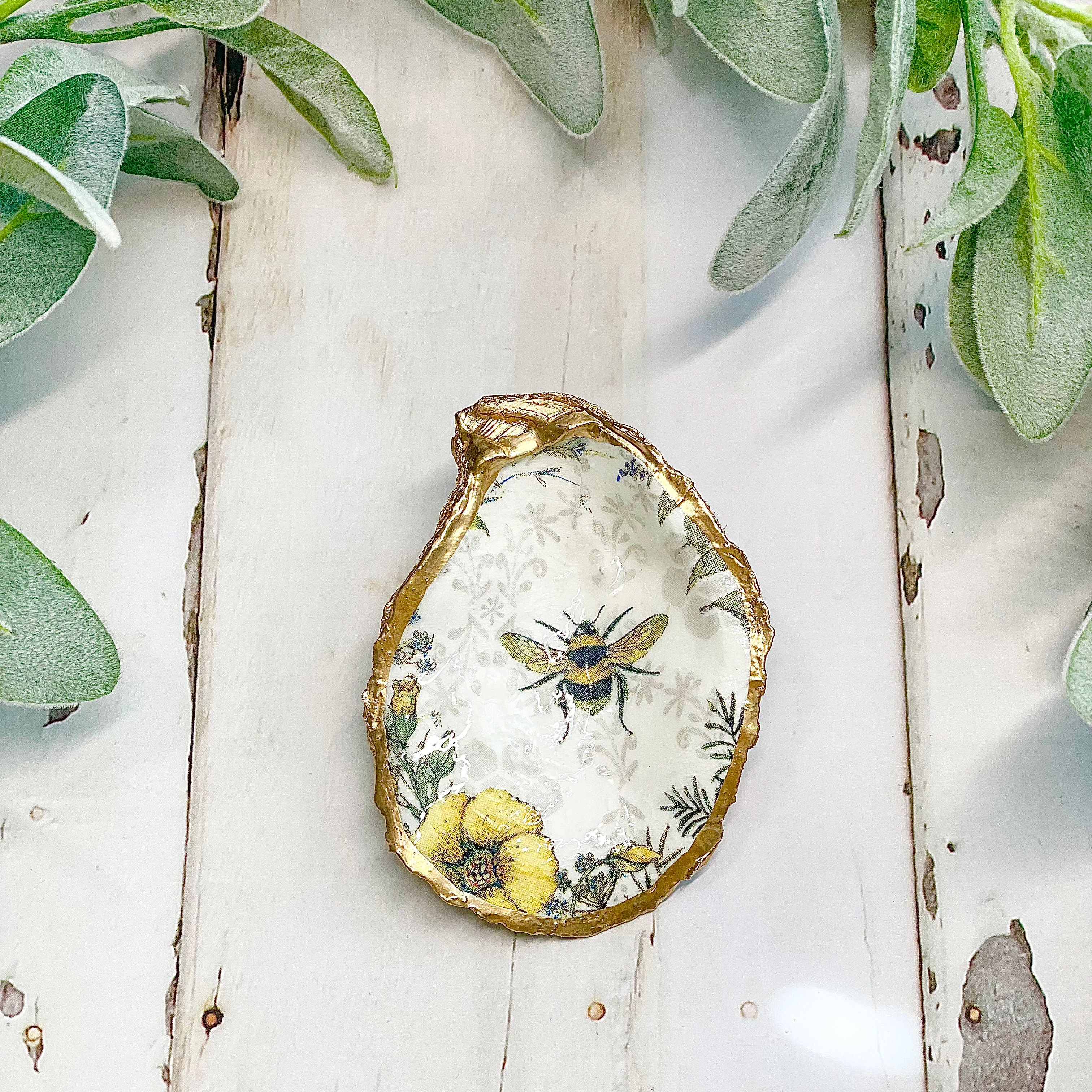 Decorative dish with a bee and floral design on a white wooden surface with green leaves.