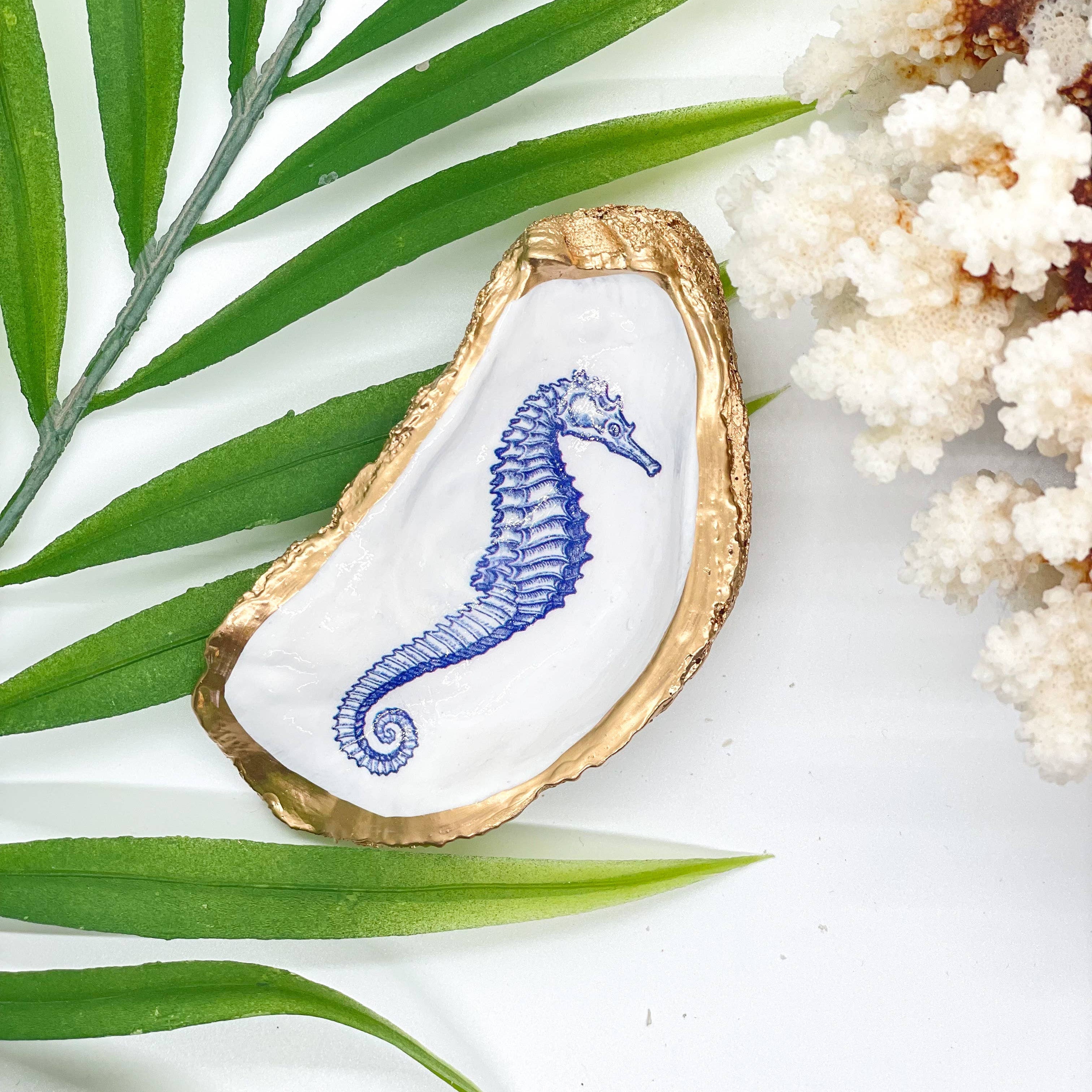 Decorative oyster shell with a blue seahorse design on a white surface with green leaves and white flowers.