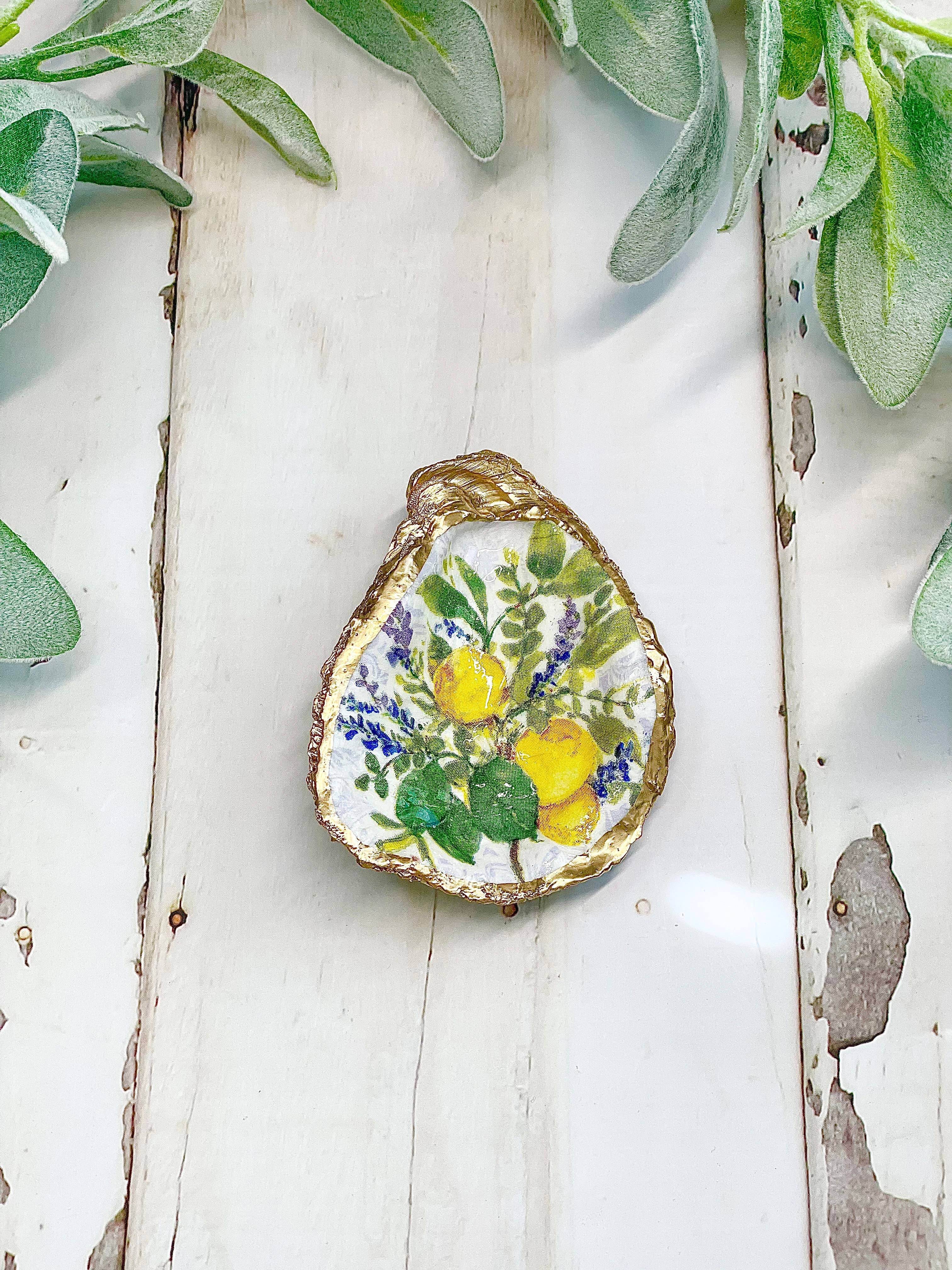 Decorative oyster shell with floral design on a white wooden surface with green leaves.